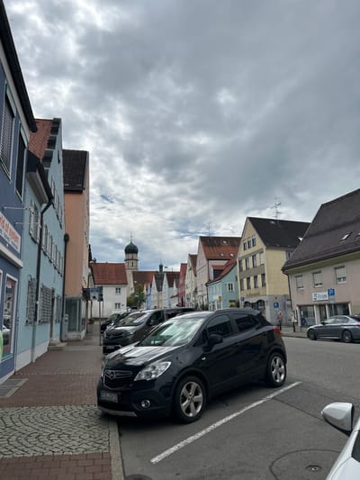 The image depicts an overcast daytime street scene in Schongau, Germany. The view is oriented down a paved thoroughfare, with a prominent church steeple featuring an onion-shaped dome visible in the distant background.

The sky is uniformly covered with dense, grey clouds. The street surface consists of asphalt with white painted lines delineating parking spaces. The sidewalk on the left is paved with reddish brick pavers transitioning to a cobblestone pattern.

Multi-story buildings with varied facades and pitched gable roofs line both sides of the street. These structures are painted in pastel shades including light blue, pink, yellow, and beige. The building in the immediate left foreground is blue with exterior roller shutters on its windows and a partial sign displaying "world of system" and "Trank, Multimedia." Further down on the right, a yellow building displays a "Volksbank" sign, and an adjacent building has an "S-Team" sign.

Multiple vehicles are parked on the street. In the foreground, a black Opel Mokka compact SUV is parked facing the viewer's left, displaying a German license plate "SOG DN 15." Directly behind it is a dark-colored minivan. Further down the street on the right, a dark-colored sedan is parked. The side mirror of a white vehicle is partially visible in the extreme right foreground.

One individual is visible on the right-hand sidewalk, appearing to be an adult male wearing a red top, standing near the yellow Volksbank building. No other individuals are clearly visible or engaged in distinct actions. Streetlights, rooftop antennas, and parking signs are also present within the scene.