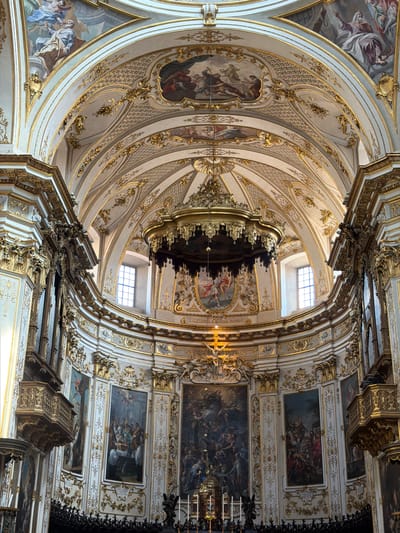 This is a photograph of the interior of a church, likely the Duomo di Mozzo, located in Mozzo, Italy. The image showcases the opulent baroque architecture of the ceiling and apse. The ceiling is intricately adorned with frescoes and gilded decorative elements, featuring detailed stucco work and ornate gold trim along the arched vaults. A large, elaborate chandelier hangs from the central dome. The walls of the apse are decorated with large oil paintings depicting biblical scenes, framed by gilded architectural details. Two large pipe organs are visible on either side of the apse, their ornate casings made of dark wood and gilded elements. Numerous windows with arched tops are present in the upper sections of the apse, allowing natural light to filter into the space. The overall impression is one of grandeur and religious devotion, typical of historical Italian cathedrals.