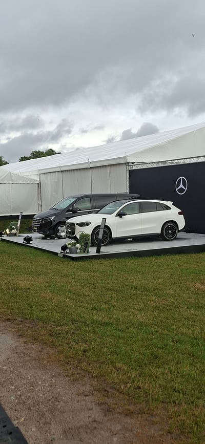 Two vehicles, a black van and a white SUV, are displayed on a raised platform in front of a large white tent structure. The van is positioned to the left of the SUV. The SUV is a Mercedes-Benz, indicated by the logo on the black backdrop behind it. The backdrop also features a large Mercedes-Benz logo. The scene is outdoors, with green grass in the foreground and a cloudy sky overhead. There are no people visible in the image. The environment suggests an outdoor event or exhibition. The specific location in Kuressaare, Estonia cannot be determined from the image itself.