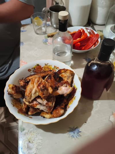 In Il'mino, Russia, a table is laden with food and drinks. A bowl of grilled chicken wings sits prominently, its golden-brown skin glistening. Beside it, a bowl of sliced red bell peppers offers a vibrant contrast. Two bottles, one containing a clear liquid and the other a dark reddish beverage, stand ready. A glass with a slice of lemon and a metal mug are also present. The table is covered with a patterned tablecloth, and a person's arm is visible on the left side of the frame. The scene suggests a meal being prepared or enjoyed.