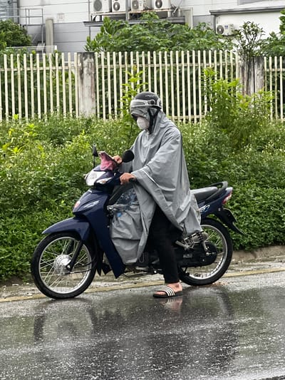 A person wearing a grey poncho, white mask, helmet, and sandals rides a blue motorcycle on a wet street. The person is on the left side of the frame. The motorcycle is facing towards the right. The background consists of a white fence, green bushes and trees, and a building with air conditioning units. The street is reflective due to the rain. The location is Sơn Dương Commune, Vietnam.
