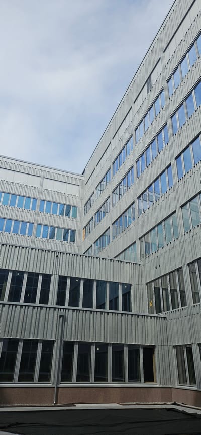The image shows a modern, multi-story building with a facade of metal paneling and numerous rectangular windows. The building appears to be made of light-colored metal, possibly aluminum or steel, with a ridged texture. The windows are predominantly dark, reflecting the sky, with some showing a blue hue, suggesting they are tinted or the sky is clear.
The building is angled in the shot, with the bottom and right sides of the image showing more of the structure while the top and left sides are dominated by a light blue sky, indicating a bright, possibly daytime setting. There is a sense of architectural design with a combination of solid panels and glass sections, creating a rhythmic pattern across the facade. Some sections of the building have a more pronounced grid-like appearance, while others feature longer, vertical window arrangements.
In one section, a yellow "X" mark is visible on the metal facade, possibly indicating construction or maintenance work. There is no indication of people or any specific activity taking place; the scene appears to be a static architectural study. The overall impression is one of contemporary design and urban architecture. Given the location context of Västerås, Sweden, this could be an office building, a residential complex, or a public institution. The lighting suggests it is daytime, and the clear sky points to fair weather.