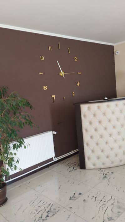 The image depicts a reception area, likely in a hotel or business in Khmilnyk, Ukraine. A large, modern wall clock with gold numerals and hands dominates the brown wall. Below the clock, a white radiator runs along the bottom of the wall, with a potted green plant to its left. To the right of the clock and radiator, a tufted, cream-colored reception desk is partially visible. The floor is tiled with a light-colored marble pattern. A security camera is mounted high on the right wall. No people are visible in the scene.