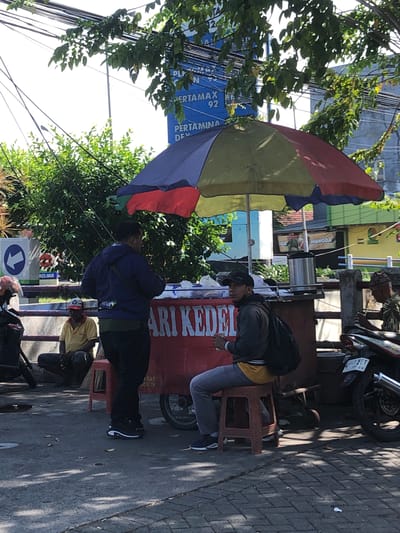 In Banjarkemantren, Indonesia, a street scene unfolds under a bright sky, with dappled sunlight filtering through overhead trees and power lines. A colorful umbrella, segmented in red, yellow, and blue, shades a food cart adorned with large red lettering that reads "ARI KEDELAI". To the left of the cart, two men are visible. One man, wearing a dark blue jacket over a green shirt and dark pants, stands near the cart. Beside him, a man in a yellow shirt and dark pants sits on the ground, appearing to be resting. To the right of the food cart, another man, dressed in a gray jacket over a yellow shirt and gray pants, sits on a red stool, holding a white object in his right hand. He wears a black baseball cap. In the background, motorbikes are parked, including one with a license plate that reads "2443 NET 01-29". Buildings with yellow and green facades are visible further in the background, along with some foliage. A blue sign with a white arrow pointing left is also present on the left side of the image.