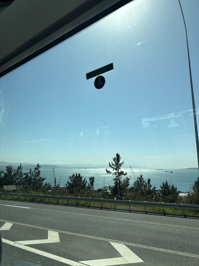 The image captures a sunny daytime view from inside a vehicle, looking out at a coastal highway and a sparkling body of water. The scene is set in Dilovası, Turkey.
In the immediate foreground on the left, the edge of a vehicle's windshield and interior frame are visible. A dark sticker with a circular design and a rectangular bar is affixed to the windshield, catching sunlight. The windshield also shows reflections of what appear to be interior structures or external light sources.
To the right, a multi-lane asphalt highway stretches diagonally, marked with clear white lines, including a large, partially visible numeral "7" or an arrow. A metal guardrail separates the highway from a narrow strip of green grass and a line of dark green, coniferous-like trees.
Beyond the trees, a vast body of water, likely the Gulf of Izmit, glistens intensely under the bright sun, creating a shimmering effect. Several large ships or cargo vessels are visible in the distance across the water. The horizon features hazy, distant land or mountains, indicating a wide expanse. Some industrial cranes or structures are faintly discernible along the distant shore. The sky above is a clear, light blue, with a strong sun flare evident in the upper left quadrant of the windshield.
The overall impression is of a scenic drive along a coastal road on a clear, bright day. No distinct text is readable in the image.