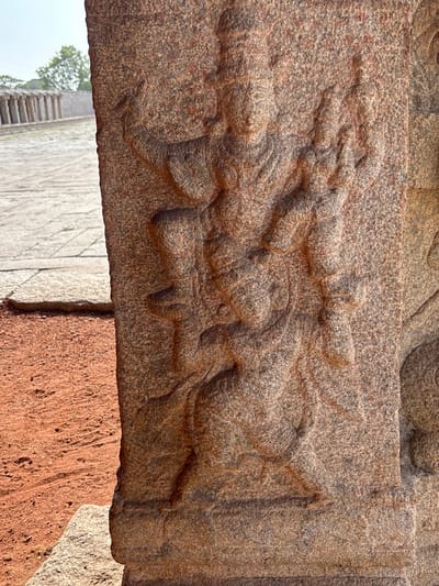 The image shows a close-up of a stone carving, likely part of an architectural structure in Kamalapura, India. The carving depicts a mythical creature, possibly a Yali or similar composite animal, rendered in relief on a rectangular section of brownish-gray stone. The creature has a textured surface, with details such as facial features, limbs, and possibly wings or decorative elements suggested by the carving. The surrounding stone surface is rough and weathered, suggesting age and exposure to the elements. The carving is positioned horizontally, and a section of another stone structure or wall is visible beneath it, showing a transition to a different color and texture of stone. In the background, faint outlines of what appear to be steps and vegetation can be discerned.