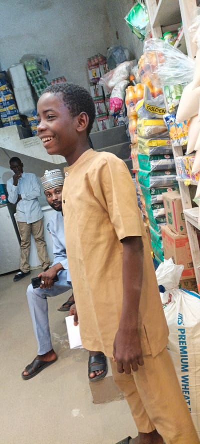 The scene depicts an interior view of a commercial establishment, likely a shop or storage room, in Daurawa, Nigeria.
**People:**
1. **Foreground:** A young male, approximately 8-12 years old, with dark skin and short, dark curly hair, stands facing right but looking slightly left out of frame. He wears a light brown (tan/khaki) short-sleeved collared shirt with two black buttons on the placket and matching trousers. His right arm is largely obscured, while his left arm is visible, hanging loosely. He is smiling broadly, exposing upper teeth. He wears dark-colored open-toed sandals.
2. **Mid-ground:** A male individual with a beard and a light-colored, patterned cap is seated on what appears to be a low surface or stool behind the smiling boy. He wears a light blue long-sleeved shirt and light blue trousers, along with dark sandals. He is looking directly towards the viewer. His right hand holds a dark smartphone, and his left hand holds a small white piece of paper.
3. **Background:** Another male individual stands on the left side of the frame, partially obscured by the foreground subjects. He has dark skin and wears a light blue long-sleeved shirt and light brown (tan/khaki) trousers. His right hand is raised towards his mouth or chin, and he is looking towards the left. He wears dark shoes.
**Objects and Environment:**
The room features light-colored walls, possibly concrete or plaster. The floor is also light-colored. The background is filled with various merchandise and storage items.
* **Right Side:** Multiple shelves are visible, densely packed with products. These include stacks of packaged goods in plastic, plastic bottles of yellow liquid (possibly juice or oil), bags labeled "GOLDEN," and numerous other small packaged items, some in transparent plastic wrapping. A large white sack labeled "PREMIUM WHEAT" is visible towards the bottom right. Cardboard boxes are also stacked.
* **Left Side:** Stacks of merchandise in boxes and plastic wrapping are piled against the wall. A tall, white appliance, possibly a refrigerator or freezer, is partially visible behind the standing man.
**Actions and Interactions:**
The young boy is smiling widely, looking past the viewer's left. The seated man is observing towards the viewer while holding a smartphone and paper. The standing man appears to be in a contemplative pose. No direct interaction between the individuals is actively depicted.