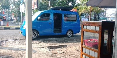 The scene depicts a daytime roadside environment in Bojongherang, Indonesia. In the foreground on the right, a wooden street vendor cart with glass panels is visible, containing a red plastic basket filled with numerous white, spherical food items. An off-white vertical support pole stands partially on the extreme right, with a grey umbrella or awning extending above. In the midground, a bright blue passenger van, equipped with a black rooftop cargo box and aftermarket silver multi-spoke wheels, is parked on the asphalt shoulder of a road. Its passenger-side sliding door is open. The ground directly in front of the viewer consists of uneven, cracked dirt. To the left and in the background, a striped curb, an orange traffic cone, various parked and moving vehicles (including motorcycles), trees, and commercial signage (one featuring "SMBC" text) are present. No identifiable human subjects are discernible.