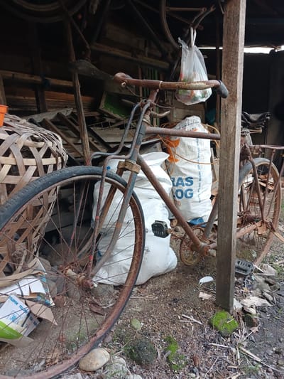 The image depicts a cluttered, rustic scene, likely an outdoor storage area or shed in Geudonggeudong, Indonesia. The primary subject is an old, rusty bicycle, angled diagonally across the frame. Its frame, spokes, and wheels show significant signs of wear and corrosion, suggesting it has been left outdoors for an extended period.

Several objects are scattered around the bicycle. These include white plastic sacks, one with blue text partially visible. There's also a woven bamboo basket, possibly a pannier or storage container, positioned above the rear wheel of the bicycle. Other items include pieces of wood, rocks, and some greenery, including moss growing on a rock. The ground appears to be dirt and debris. In the background, wooden beams and planks suggest the structure of a shed or shelter. A partially visible bicycle wheel with spokes can be seen in the upper right corner.

The lighting suggests it might be daytime, but the interior of the shed is dark, with light filtering in from the right side. There is no visible weather activity. The overall impression is one of neglect and disuse.

Visible text on the white sack includes "BULOG" and part of "RAS". Another sack in the background has partial text that appears to be from "TELEPON". The context of "BULOG" and "TELEPON" suggests these are likely commodity or telecommunications-related sacks, common in Indonesia.