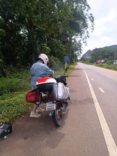 A person wearing a denim jacket and a white helmet is standing next to a motorcycle parked on the shoulder of a road in Khao Khram, Thailand. The motorcycle is loaded with a red bag and a silver-wrapped item strapped to the back. The motorcycle's license plate reads "102 สุราษฎร์ธานี 9553." The road is asphalt with a white line marking the edge. Lush green vegetation is present on the side of the road where the motorcycle is parked, while the opposite side of the road features more sparse vegetation and buildings in the distance. A blue and yellow sign is visible near the person and motorcycle. A helmet is on the shoulder of the road next to the rear tire. A mountain range is in the background.
