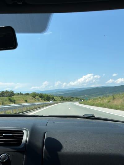 The image presents a first-person view from inside a vehicle, looking out through the front windshield onto a highway under a clear, bright sky. The scene depicts a daytime drive through a scenic, hilly, or mountainous landscape, consistent with the Blatino, Bulgaria region.
In the lower right portion of the frame, the dark gray, textured dashboard of the vehicle is visible, featuring an air vent and part of a control dial. A windshield wiper arm mechanism is situated near the center of the dashboard's top edge, and a portion of a rearview mirror is seen in the lower left.
Beyond the windshield, an asphalt highway stretches into the distance, gently curving to the right. White lane markings, including a solid white line on the right shoulder and dashed lines, are clearly visible. A metal guardrail borders the right side of the road. Further along the road, a small, light-colored vehicle is discernible.
The landscape flanking the highway consists of green hills and mountains covered with trees and vegetation, extending under a wide, blue sky. Scattered white cumulus clouds drift across the sky, and a very small, faint object, possibly an airplane, is visible high up. The clear conditions and bright sunlight suggest good weather for travel. The overall impression is one of tranquil movement through a natural, open environment.