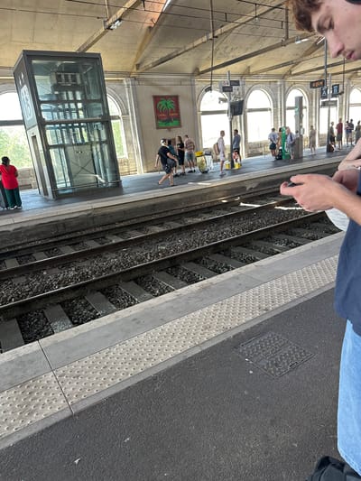 This media file captures a scene on a train platform, likely in Nîmes, France, given the context. The perspective is from a person standing on the platform, looking down at the train tracks. The platform itself is a concrete structure with a textured, tactile warning strip along the edge closest to the tracks. A set of train tracks, filled with gravel, runs parallel to the platform.

Several people are visible on the platform, some waiting, others moving. They are dressed in casual summer clothing, suggesting warm weather. In the background, the station architecture is visible, featuring arched windows and a covered roof structure supported by columns. A modern glass-enclosed elevator or lift is also present on the platform, indicating accessibility features.

Above the people and further down the platform, a framed picture hangs on the wall, depicting a palm-like plant with the text "COL NEM" visible. A digital clock on the platform displays the time as "16 05 23", suggesting it's late afternoon. A dark manhole cover with embossed text is visible on the paved area of the platform, but the text is not fully legible. The overall atmosphere appears to be that of a typical operational train station on a bright, sunny day.