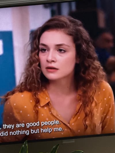 The image shows a young woman with curly brown hair and a polka-dot orange shirt. She is looking to her left with a concerned expression. There are subtitles at the bottom of the image that read ", they are good people. did nothing but help me." The background is blurred, but it appears to be an indoor setting with some blue and pink elements on the left and darker figures on the right. The image is a screenshot of a screen, possibly a television or computer monitor. The location of Springboro, United States of America is not discernible from the image.