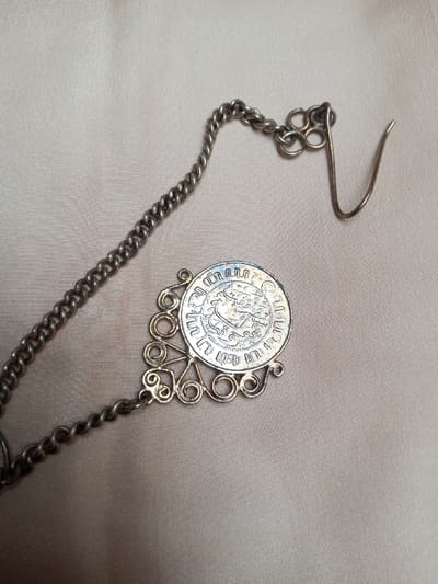 A single piece of tarnished silver-colored metal jewelry is depicted resting on a smooth, light beige fabric surface. The jewelry comprises a fine curb-link chain, a circular medallion, a filigree decorative element, and an ear wire hook. The chain spans diagonally from the upper left to the lower right of the frame. The circular medallion, positioned towards the lower right, features an embossed design of a stylized figure surrounded by script or symbols along its rim, with visible blueish discoloration on its surface. Attached to the medallion is a semi-circular filigree component, fashioned from scrolled wirework, connecting it to the chain. At the upper right terminus of the chain, a simple, bent wire hook, characteristic of an earring finding, is present. All metallic components exhibit significant tarnish and surface wear. The background fabric displays a subtle sheen and soft folds. The lighting is diffused. This scene is located in Qarabucaq qəsəbə, Azerbaijan.