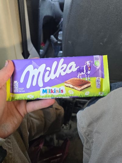 A light-skinned human hand holds a Milka Milkinis chocolate bar in the interior of a vehicle, in the city of Opalchenets, Bulgaria. The chocolate bar packaging is primarily purple with yellow borders, displaying the white "Milka" logo, an illustration of a purple and white Alpine cow, and a depiction of a chocolate-filled wafer bar. Text on the packaging indicates "100% ALPINE MILK" and "SUSTAINABLY SOURCED COCOA." The background features the dark grey fabric of a vehicle seat and a lighter-colored interior door frame. The hand is positioned towards the lower left of the frame, holding the bar horizontally across the center.