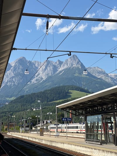 A train station platform runs alongside a train with numbered carriages. Overhead wires stretch across the sky, with mountains in the background. A sign reading '150' is visible on one of the wires. The platform is partially covered by a roof. The view looks towards a forested hillside below the mountains.