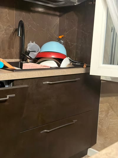 The image shows a cluttered kitchen sink area in a residential dwelling located in Spinea, Italy. A black faucet is positioned above a dark-colored sink filled with dirty dishes, including bowls, plates, and a light blue pot. A pink dish towel is also visible in the sink. The backsplash behind the sink is a brown marbled tile. To the right of the sink, a white framed window with sheer curtains is partially visible. Below the sink are dark brown cabinets with silver handles. The overall impression is one of a domestic scene where cleaning tasks are pending.
