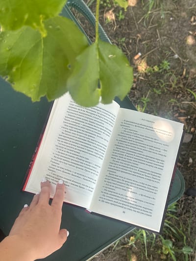An open book lies on a green surface, possibly a table or chair, with green leaves partially obscuring the scene from above. A person's hand with white nail polish rests on the left page of the book, indicating an interaction with the reading material. The book is open to pages with Ukrainian text. The surrounding environment appears to be outdoors, with dirt and grass visible in the background, suggesting a natural or park-like setting. The overall impression is one of a relaxed, possibly solitary, moment of reading in an outdoor environment. The image does not contain enough information to pinpoint a specific location within Yablunivka, Ukraine, as it lacks any distinguishing landmarks or street signs.