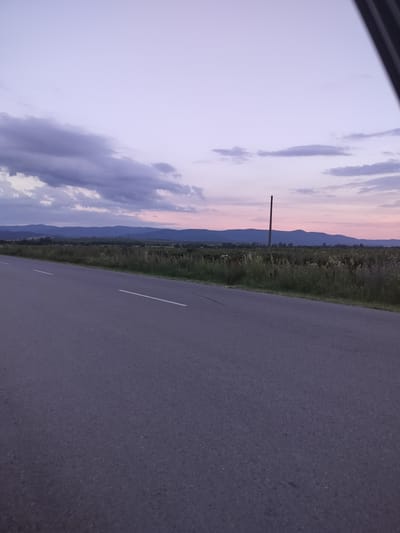 A paved road with white dividing lines extends into the distance, angling from the lower left to the middle right of the frame. The road surface is a dark grey asphalt. To the right of the road is a field of green grass and wildflowers, with some taller weeds. In the background, a line of dark blue hills stretches across the horizon. A single, thin, dark pole stands vertically in the middle of the field. The sky above is a gradient of soft purple and pink, with some scattered grey clouds in the upper left. The scene appears to be located in or around the village of Manolovo, Bulgaria, possibly near a rural road leading into the countryside.