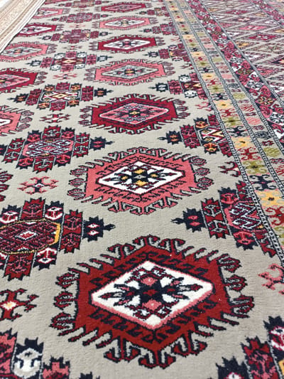The image displays a close-up, angled view of a traditional rug with intricate geometric patterns. The rug features a beige background adorned with repeating motifs in shades of red, pink, black, and yellow. Central to many of the patterns are diamond shapes containing smaller, stylized designs. The overall design suggests a handcrafted item, possibly a carpet or prayer rug. The texture of the rug fibers is visible, indicating a plush surface. The lighting appears to be natural and diffused, casting no harsh shadows. The setting is neutral, with no environmental cues other than the rug itself. There is no visible text in the image. The perspective draws the viewer's eye along the length of the rug, emphasizing its repeating design and the detailed craftsmanship.