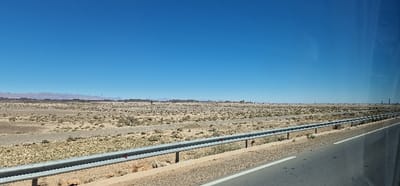 The image depicts a vast, arid landscape under a clear blue sky, identified as Ferkla El Oulia, Morocco. In the immediate foreground, the right side features a paved road with a solid white edge line and a broken white center line, suggesting a travel lane. To the left of the road, a galvanized steel guardrail runs horizontally across the lower portion of the frame, supported by multiple dark posts. Beyond the guardrail, the terrain is predominantly flat, sandy, and light brown, dotted with sparse, low-lying scrub vegetation. An unpaved, gravelly track or dry riverbed crosses the midground from left to right. In the far background, a long, low range of dark, jagged mountains stretches across the horizon. Below these mountains and extending into the midground, numerous rectangular, dark structures are visible, possibly comprising a solar energy field or industrial complex. Several slender, dark vertical structures, resembling utility poles or towers, are scattered across the distant landscape. No individuals or active subjects are present. The scene is brightly illuminated by direct sunlight.