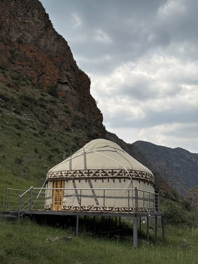 The image depicts a traditional Kyrgyz yurt situated on a raised platform in a mountainous landscape. The yurt, made of canvas and supported by a wooden frame, is decorated with patterned trim. A metal railing surrounds the platform, which has stairs leading down to the grassy ground. The surrounding environment consists of steep, rocky slopes covered in sparse vegetation and a cloudy sky overhead. In the background, distant mountains are visible. The scene is serene and appears to be in a natural, undeveloped area. The location is identified as the city of Kyzyl-Korgon, Kyrgyzstan.