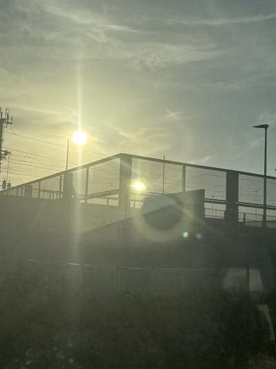 The image depicts a bright, hazy sun shining through a sky with wispy clouds. The sun's rays create a lens flare effect, and a distinct halo is visible around one of the sunbeams, suggesting it might be a double sun or a reflection. In the foreground, a metallic fence-like structure, possibly part of a building or a stadium, is visible. The structure has a grid pattern and appears to be made of dark metal. Reflected light and some green and white dots are visible on the surface, possibly from the fence or a window. Below the fence, there are some darker, indistinct shapes that could be foliage or other objects. At the very bottom of the image, a portion of a utility pole with wires and some mechanical components is visible, indicating an outdoor, possibly urban or semi-urban, setting. The overall lighting suggests it is either sunrise or sunset, with the sun low in the sky. The presence of the fence and utility pole provides some context for the location. The image is taken from a slightly elevated or angled perspective, looking upwards towards the sun and the structure. There is no visible text in the image.