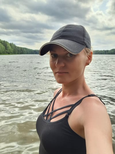 In an outdoor medium shot, a fair-skinned woman with blonde hair, wearing a black baseball cap and a black tank top with a caged neckline, looks directly at the camera. She is positioned in the foreground, with a body of water and a treeline in the background under a cloudy sky. The location is the lake in Novovilgovskoye Sel'skoye Poseleniye, Russia. The water is rippling, and the trees form a dense, dark green wall along the far bank of the lake. The sky is overcast with large, grey clouds.