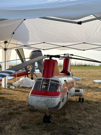 A model helicopter is situated on dry grass under a white canopy. The helicopter is red and white with black rotor blades. A person is visible in the background, wearing light-colored pants and a light-colored shirt. Another model airplane is partially visible to the left of the helicopter. The scene appears to be an outdoor event or gathering. The location is identified as Becherbach, Germany.