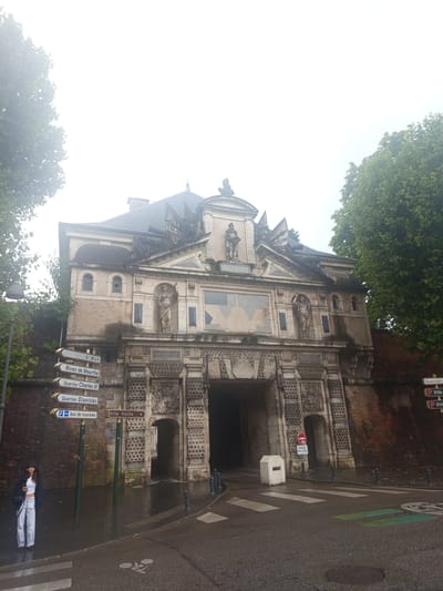 The image shows the Porte de la Craffe, a historic city gate in Nancy, France. The gate is a large, stone structure with intricate carvings and statues. It is flanked by brick walls and mature trees. The sky is overcast, and the ground is wet, suggesting it has recently rained.
In the foreground, to the left of the gate, a young woman stands on the sidewalk, wearing a hooded jacket, white top, and light-colored pants. She is holding a cup in her left hand and a phone in her right. Several directional signs are visible on poles in front of the gate, pointing to various locations within Nancy, including "Rives de Meurthe," "Quartier Charles III," "Quartier Stanislas," and "Bus de tourisme."
To the right of the gate, a street with a crosswalk is visible. There are also bollards and a white trash receptacle. A "Ville Vieille" (Old Town) sign is also present. Further to the right, on the wall, a parking sign is partially visible. The overall impression is of a historical site in a city that experiences regular rainfall.