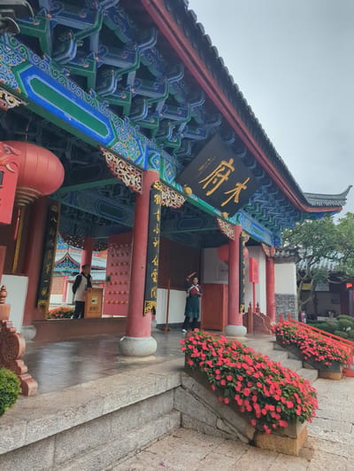 The image captures the entrance to a traditional Chinese building in Gucheng District, China. The building features a brightly painted roof with intricate blue, green, and red decorative patterns. A large black sign with golden Chinese characters is prominently displayed above the entrance. Red lanterns hang on either side of the entrance, and red pillars support the eaves of the roof.
Two individuals are visible at the entrance. On the left, a person in a light-colored jacket stands near a ticket check sign. On the right, a person in a dark green outfit is posing with their hand on their head, looking towards the left.
The foreground is adorned with planters filled with vibrant red flowers, cascading down the stone steps leading to the building. The ground is paved with stone, and parts of it appear wet, reflecting the overcast sky. In the background, more traditional Chinese architecture is visible, along with lush green trees and foliage. The overall scene conveys a sense of cultural heritage and natural beauty.