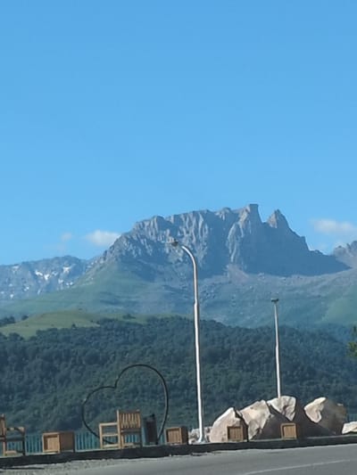 This image depicts a scenic view of a mountain range under a clear blue sky, likely captured from a roadside vantage point in or near Yeni Zod, Azerbaijan. In the foreground, a dark metal heart-shaped sculpture is prominent, with several wooden chairs and small wooden boxes placed behind a railing. Two modern streetlights stand nearby, one on the left and another further to the right. The middle ground features a dense, dark green forest covering rolling hills. The background is dominated by a majestic, rocky mountain peak, with some snow visible on the higher slopes. The overall impression is one of natural beauty and potentially a public recreational area.