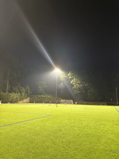 This image captures a nighttime scene at an illuminated artificial turf sports field in Newton, United States. A tall floodlight pole stands centrally, emitting intense white light that forms a visible beam upwards into the dark sky and brightly illuminates the playing surface. The field features green artificial turf with white and blue demarcation lines. In the background, dense green trees line the perimeter, with a section of tiered concrete bleachers visible behind them. Approximately seven individuals are present on the field; most wear purple athletic shirts, while one on the far left wears a yellow shirt. Two soccer goals with nets are visible, one in the mid-ground right and another partially on the far left. The individuals are dispersed across the field, indicative of ongoing athletic activity, likely a soccer game or practice under artificial light.