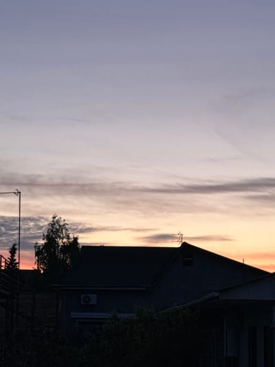 The image depicts a dusk or dawn sky in Береговой сельский совет, Ukraine, with pastel colors transitioning from pale orange to muted purple. Wispy clouds stretch across the sky. In the lower right, a darkened building is visible, featuring a sharply angled roof. An air conditioning unit is mounted on the building's exterior. A tree with sparse foliage sits in front of the building, obscuring some of the structure. An electrical pole is situated near the bottom center of the image. The scene appears still and quiet, suggesting a residential or suburban location.