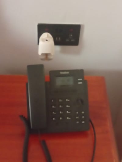 A black desk phone with a handset rests on a reddish-brown table. The phone features a digital display and a standard numerical keypad. Above the phone, mounted on a light-colored wall, is a black electrical outlet. Plugged into the outlet is a white air freshener. The overall scene appears to be in a hotel room.