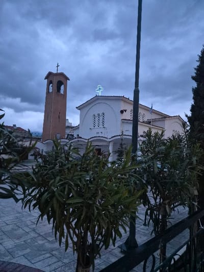 This image depicts the Church of Agios Nikolaos (Saint Nicholas) in the Municipal Unit of Corinth, Greece, under an overcast sky. The prominent subject is a white-painted Greek Orthodox church building with a distinctive facade featuring arched windows with decorative grilles and a blue neon cross illuminated on its roofline. A Greek flag is visible flying from a pole on the church's right side. To the left of the main church stands a reddish-brown brick or stucco bell tower with arched openings, also topped with a cross.

In the foreground, several green shrubs with long, narrow leaves are visible, partially obscuring the lower portion of the scene. A dark metal railing runs diagonally from the lower right. The ground is paved with light-colored rectangular pavers, forming a plaza or courtyard leading up to the church. A tall, dark utility pole stands vertically in the mid-ground, positioned in front of the church's right side. The sky is filled with dark grey-blue clouds, indicating dim lighting, possibly dusk or an overcast day. No individuals or discernible actions are present in the scene.