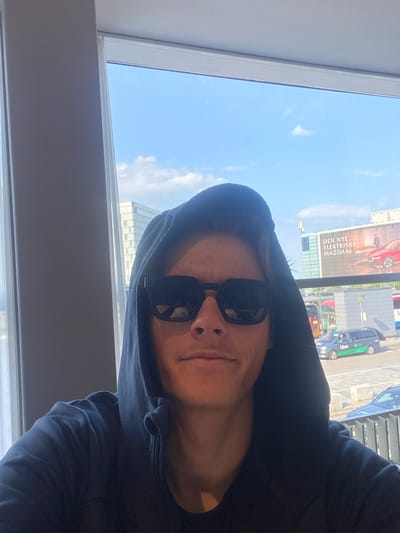 The image is a selfie taken from a low angle, showing a person wearing sunglasses and a hooded sweatshirt. The person is indoors, near a window. Through the window, there is a view of Kastrup, Denmark. Specifically, a large billboard is visible advertising a Mazda 6e, along with street-level views including a green-accented Uber vehicle. The sky is blue with some clouds.