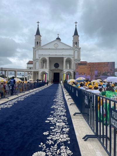 A grand white church with two prominent bell towers topped with crosses stands under an overcast, cloudy sky. The church features a classical facade with arched entrances and columns. Leading up to the central entrance is a long, navy blue carpet adorned with intricate white floral patterns along its borders.

The carpet is flanked by blue metal crowd control barriers. Behind these barriers, a crowd of people is gathered, many wearing yellow and white attire. Some individuals are holding open white umbrellas, and others appear to be wearing hats or caps. On the right side, a large outdoor screen displays an ornate, possibly religious, interior scene. Near the church's main entrance, several vertical flags in colors like red, yellow, and green are visible.

Visible text on the church facade reads: "A LA INMACULADA CONCEPCION DE LA VIRGEN MARIA PATRONA DE GUINEA ECUATORIAL," dedicating the church to the Immaculate Conception, Patron Saint of Equatorial Guinea. On the blue barriers and a police vest, the words "POLICIA" and "POLICIA NACIONAL" are clearly visible, indicating the presence of security forces. The scene suggests a significant public or religious event taking place during the daytime in Mongomo, Equatorial Guinea.