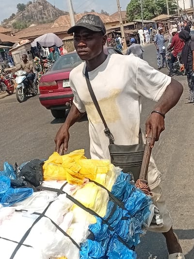 The image depicts a bustling street scene in Ozuri, Nigeria. In the foreground, a young adult male with dark complexion is standing, slightly turned to his left. He wears a black baseball cap with "D&G Exchange" in white lettering, a white short-sleeved t-shirt with a light-colored stain on the front, light-colored shorts, and white slides/sandals. A black shoulder bag with a zipper pocket is slung across his body. His right hand is near a heavily loaded pushcart, which contains numerous bundles of plastic bags in yellow, white, blue, and black, secured with black straps. His left hand holds a dark, rod-like object, possibly a stick or umbrella handle, resting against the cart.
In the midground, a red sedan is visible behind the man, with a partially obscured license plate beginning "SAN". To the left of the sedan, a person rides a silver motorcycle. Numerous other individuals, both pedestrians and motorcyclists, are present across the street. These individuals are dressed in various attire, including traditional patterned clothing. The background features several single-story and two-story buildings with corrugated metal roofs, indicative of commercial or residential structures. A prominent rocky outcrop or hill rises in the distant background under bright daylight conditions. The overall scene suggests a busy market area or urban thoroughfare.