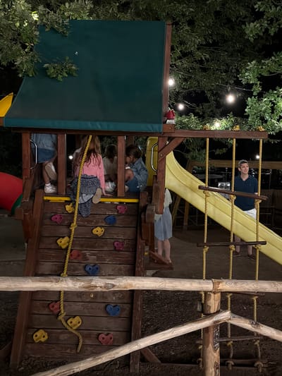 In the city of Cavriglia, Italy, a wooden and yellow plastic playground structure is illuminated by string lights at night. Several children are visible on the playground, some climbing the wooden stairs and others inside the structure. A man in a dark polo shirt and white shorts stands near the yellow slide, looking to the side. The ground around the playground is covered in mulch, and trees with green leaves are visible in the background. The scene appears to be outdoors, possibly in a park or backyard.