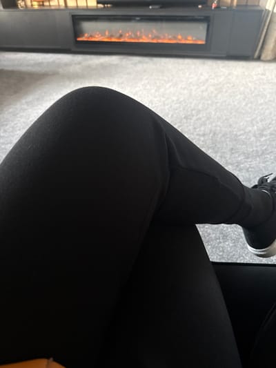 The image depicts an indoor scene in Bury, United Kingdom, focusing on the lower body of an individual seated in what appears to be a dark chair or sofa. The person's legs are crossed, with the left leg positioned over the right. They are wearing black trousers or leggings. The right foot, partially visible, is clad in a black sneaker featuring a white sole rim. The floor is covered with a light grey, textured carpet. In the background, a dark-colored entertainment unit or console table is situated against a wall. Centered within this unit is an electric fireplace displaying artificial orange flame effects.