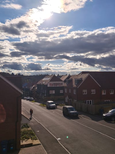 The image shows a residential street in Waverley, United Kingdom on a sunny day with scattered clouds. The street is lined with houses of varying architectural styles, primarily brick and wood-clad structures with tiled roofs. Several cars are parked along the street, including a dark-colored minivan and a silver sedan. A woman is walking on the sidewalk carrying a child. The road is paved and marked with white lines. The sky is predominantly blue with white and gray clouds, and the sun is visible, casting shadows across the scene. Trees are visible in the distance beyond the houses. Blue recycling bins stand at the edge of the sidewalk in the foreground.