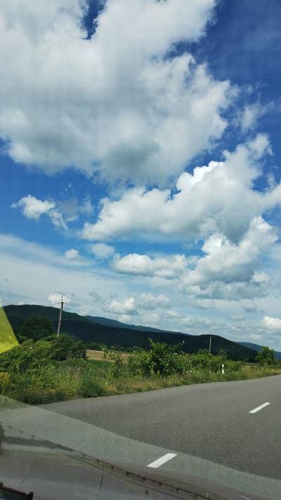 This is a landscape photograph taken from a vehicle, likely in Veliatyno, Ukraine. The image is oriented vertically.
The main subjects are the sky, a road, and a natural landscape. The sky is a prominent feature, filled with a mix of bright white and darker gray clouds against a vibrant blue backdrop. Sunlight suggests it's daytime. The road, with white lane markings, occupies the right side of the frame, indicating movement. To the left of the road, there is a lush green slope covered in trees and vegetation, with a glimpse of fields further down. In the distance, rolling hills or mountains are visible, their forms softened by the haze. A solitary utility pole with wires is present on the left side of the image.
No people are visible in the frame. The activity taking place is implied to be travel, as suggested by the road and the perspective from a moving vehicle.
Notable details include the dramatic cloud formations, suggesting potentially changing weather or a picturesque sky. The greenery points to a temperate climate, likely summer or late spring. The presence of the road and utility pole are common elements of rural or semi-rural landscapes.
There is no visible text in the image.