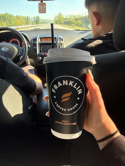 The image shows the interior of a Kia car driving on a road near Blajievo, Bulgaria. The steering wheel and dashboard are visible. A hand is holding a black Franklin House Coffee cup with a white lid and a straw. The driver is wearing black clothing and a bracelet. The rearview mirror has a tag on it. The landscape outside the window includes green trees and a blue sky. The road appears to be a highway.