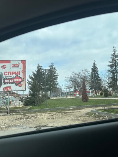 The image depicts an exterior scene in Hitrino, Bulgaria, viewed from inside a vehicle, indicated by the dark, curvilinear frame at the top and bottom edges. The sky is overcast and grey, covering approximately the top third of the frame.
In the immediate foreground, a rough, unpaved patch of ground with gravel and dirt, possibly damp, separates the viewer from the main scene. This area is bordered by a low concrete curb.
To the left of the central view, a large rectangular billboard stands on metal supports. The billboard has a red border and a white background. It features the "EDEA" logo with the slogan "Винаги добро яде!" (Always good food!). Prominent red text reads "СПРИ!" (STOP!), followed by a red arrow pointing right, containing the text "ПРИ НАС Е ПО-ЕВТИНО" (IT IS CHEAPER WITH US). Percentage symbols ("%") are scattered on the billboard, indicating discounts. A portion of a dark garment, possibly a shirt, is visible at the bottom left of the billboard.
The central and right portions of the midground are occupied by a green park area. This park features manicured grass, several tall, dark green evergreen conifer trees, and one prominent deciduous tree with bare branches. A smaller, conical evergreen shrub with reddish-brown foliage is also present. Multiple dark grey streetlights with double lamp heads are distributed throughout the park. Dirt or gravel pathways traverse the grassy areas.
In the background, behind the billboard, low-rise buildings with light-colored facades and reddish roofs are partially visible. Further back, within and beyond the park, additional structures are seen, including one with distinct red elements, possibly a commercial building, and some exposed metal framework, which could be part of construction or public amenities. No people or animals are visible in the scene, and no specific actions or interactions are depicted. The overall impression is that of a cloudy day in an urban or semi-urban environment.