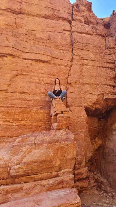 A medium shot shows a woman perched on a rock ledge within a deep, narrow slot canyon in Hevel Eilot Regional Council, Israel. The canyon walls are composed of reddish-brown rock with vertical striations and fractured surfaces. The woman is facing the camera and sitting with bent legs. She is wearing jeans, a dark tank top, and sneakers. A strap crosses her chest. The sky is visible in the upper right corner. The photograph is rotated.