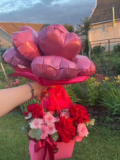 In an outdoor setting, likely a garden, a person's arm and hand are visible holding a pink box filled with a floral arrangement and a bouquet of pink heart-shaped balloons. The floral arrangement features red roses, pink roses, and small pink and white chrysanthemums, with eucalyptus leaves. The balloons are clustered at the top, obscuring much of the background which includes a tiled roof house, a fence, and green foliage. The lighting suggests it is either early morning or late afternoon, with a warm glow on the balloons and house. The location is identified as Sarotski Sel'ski Savet, Belarus.