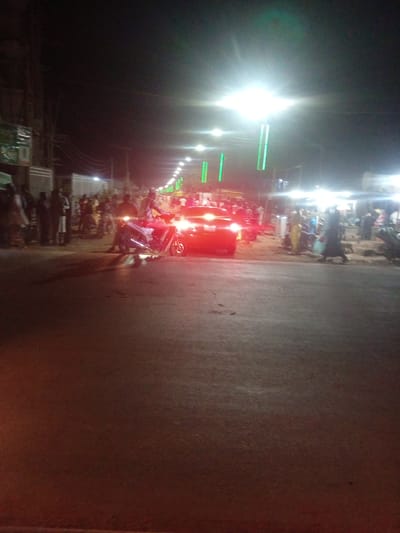 The image captures a nighttime street scene in Tundun Wada South, Nigeria, illuminated by streetlights and the headlights of vehicles. A red car is prominent in the foreground, its brake lights illuminated, suggesting it is stopped or moving slowly. To the left of the car, a motorcycle is visible with its headlight on, and a person is seated on it. A crowd of people are gathered on the side of the street, some appearing to be watching the vehicles. The background shows buildings and more streetlights, indicative of an urban environment. The overall atmosphere is one of activity on a street at night.