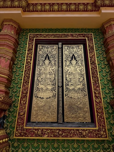 This image captures a richly decorated entrance or window of a building, likely a temple, in the city of Na Nuea, Thailand. The central focus is a pair of ornate golden doors or panels, intricately painted with detailed figures and motifs in black and gold. On each panel, a multi-armed deity stands on a lotus flower, holding various objects, with swirling decorative elements and mythical creatures like dragons below.
The doors are set within a thick, elaborately carved frame. The frame is decorated with a combination of deep red and gold, featuring floral and scrollwork patterns. Surrounding the frame is a textured wall adorned with a repeating pattern of golden motifs on a vibrant green background. The architectural elements extending beyond the frame, particularly on the left and right, are also heavily ornamented with red and gold carvings, including pillar-like structures with tiered designs. The ceiling above is painted in a warm yellow or orange hue with decorative golden trim. The overall impression is one of opulence and intricate craftsmanship characteristic of Thai religious or royal architecture.