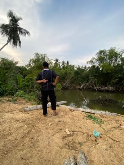 A medium shot captures a man standing on a dirt path next to a body of water, likely a canal or river, in Tanjongkapai, Indonesia. The man has his back to the camera and wears a dark, long-sleeved shirt with light-colored horizontal stripes, black pants, and sandals. He holds his left hand to his back. The vegetation surrounding the water consists of tropical trees, including palms. The sky appears overcast. The ground is dry and cracked. Along the edge of the water, logs are present. A plastic bag sits on the dirt path.
